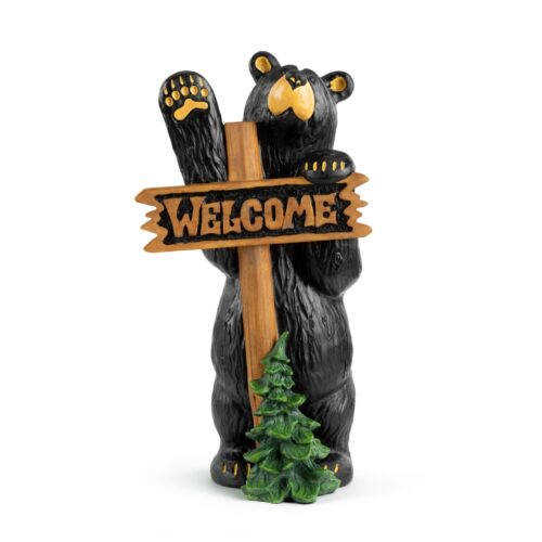 A bear holding a wooden welcome sign beside a small tree.